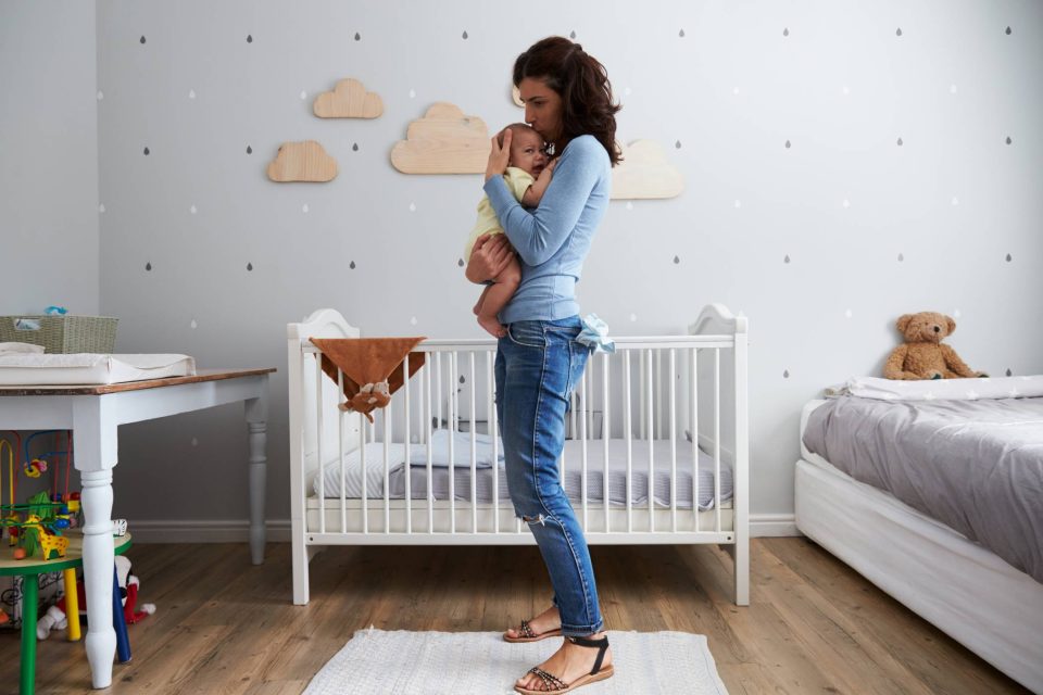 Woman standing in an infant's room with a white crib in the background, holding and soothing an infant while they cry.