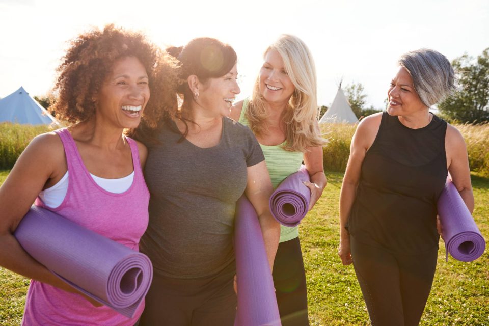 Four women of varying ages and races carrying yoga mats while walking together and smiling.