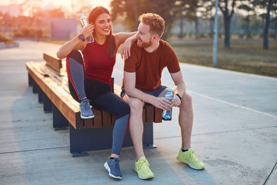 A young brunette woman and redheaded man in athletic gear sitting on a bench outside holding water bottles.