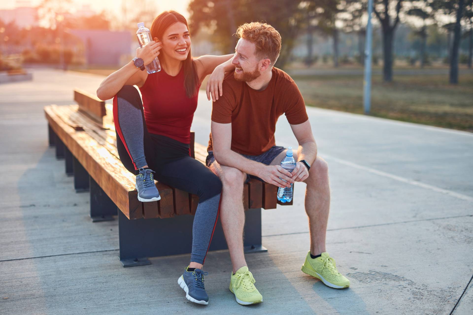 A young brunette woman and redheaded man in athletic gear sitting on a bench outside holding water bottles.