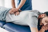 Chiropractor wearing dark blue scrubs using their hands to press against a male patient's lower back as they lay on a medical table.