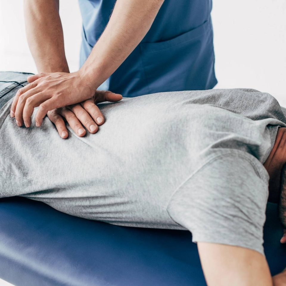 Chiropractor wearing dark blue scrubs using their hands to press against a male patient's lower back as they lay on a medical table.