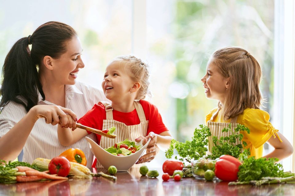 Brunette woman helping two young girls wearing aprons create a salad together.