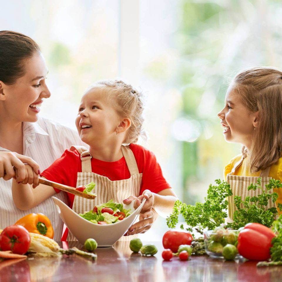 Brunette woman helping two young girls wearing aprons create a salad together.