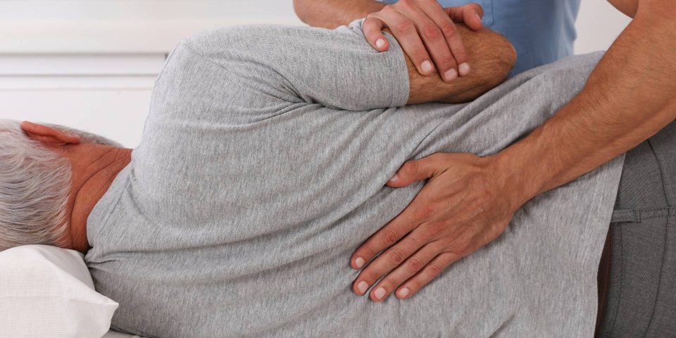 Chiropractor using their hands to gently adjust and educate an older male patient about chiropractic care in a medical office.